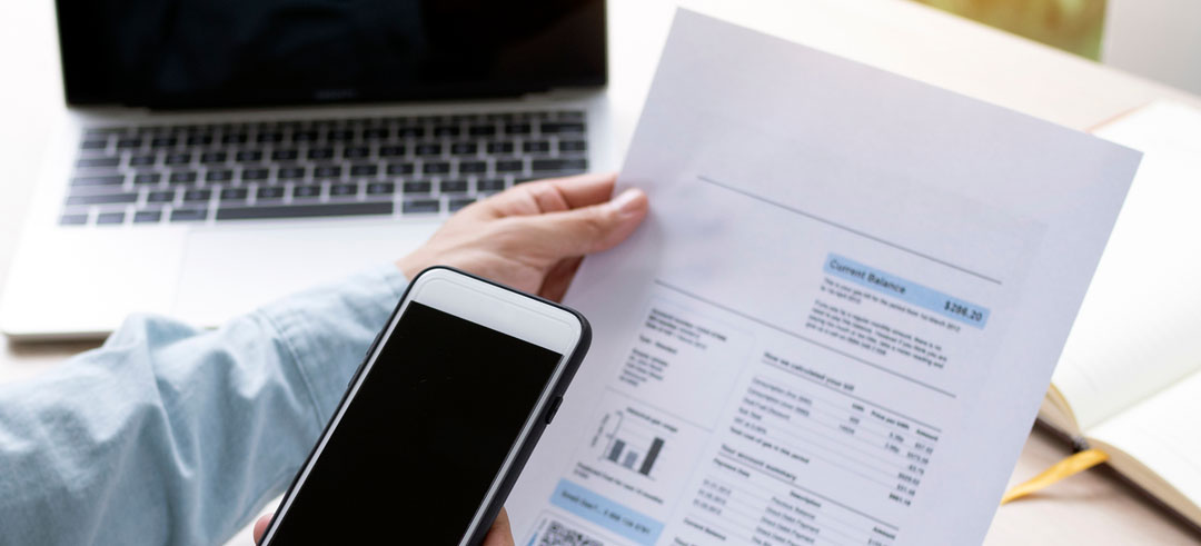 Persona sosteniendo un documento y un smartphone con un ordenador de fondo